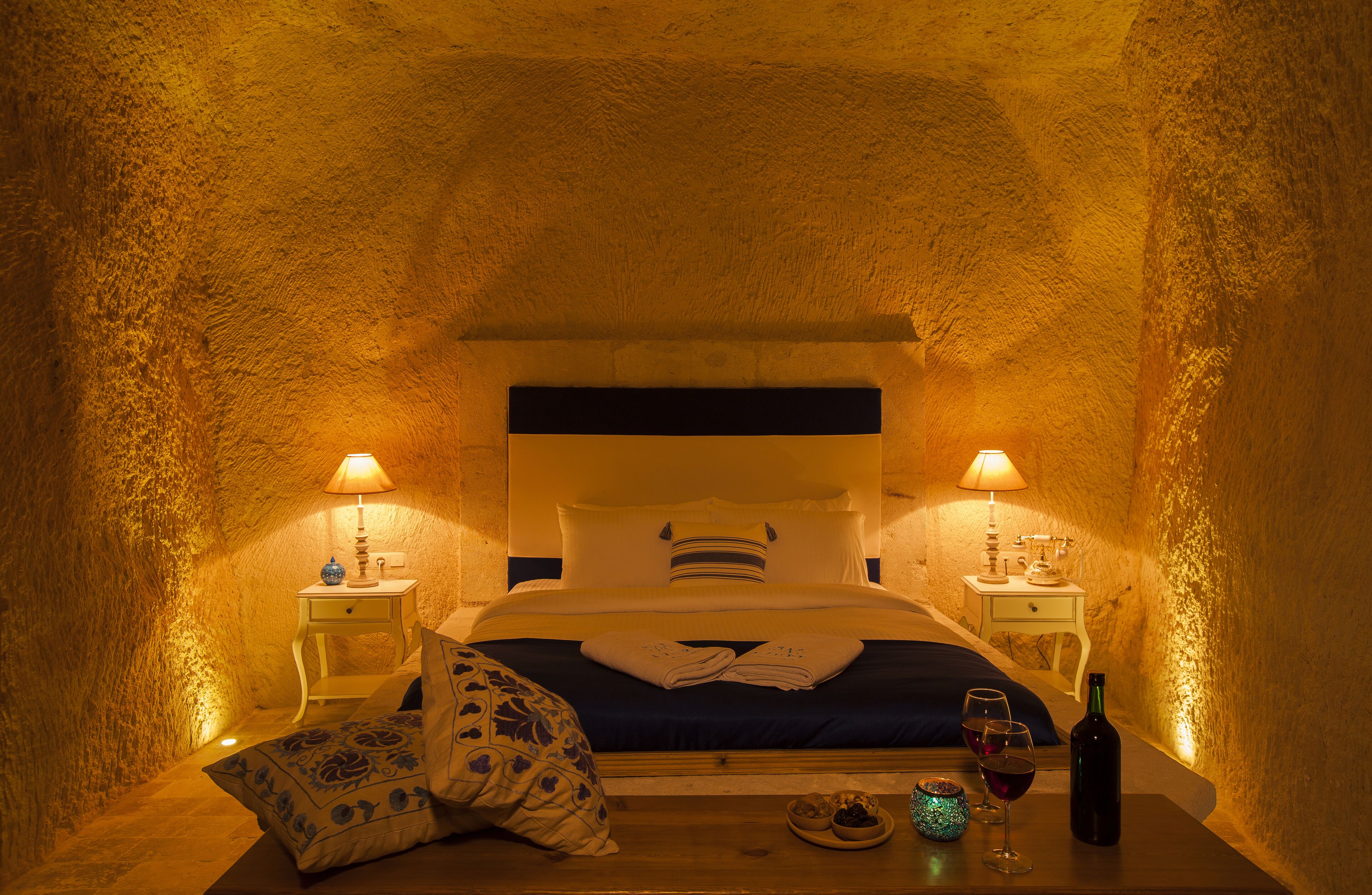 A cozy bedroom with a bed, bedside lamps, a small table with a wine bottle and glasses, and a bowl of fruit.
