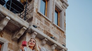 Une femme se tient sur un balcon en pierre d’un bâtiment historique et tient une tasse rouge.