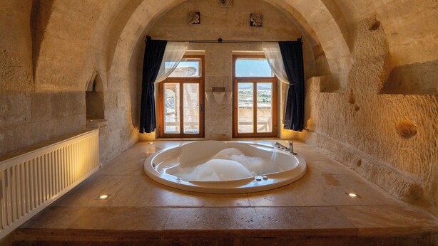 Une pièce aux murs de pierre, dotée d’une grande baignoire, d’une fenêtre avec des rideaux et offrant une vue sur l’extérieur.