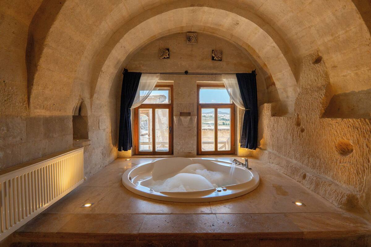A stone-walled room with a large bathtub, a window with curtains, and a view of the outdoors.