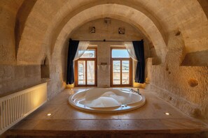 A stone-walled room with a large bathtub, a window with curtains, and a view of the outdoors. - Azure Cave Suites (Avanos)