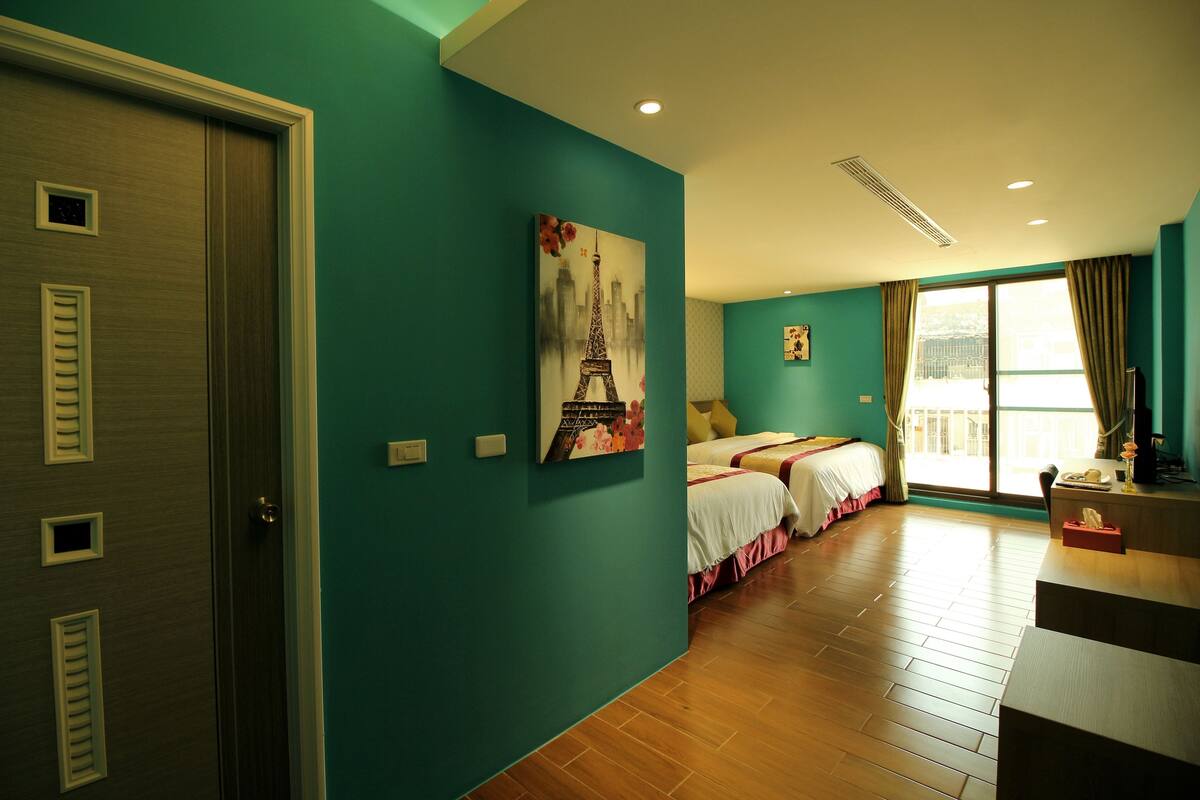 Una habitación de hotel con paredes color turquesa, piso de madera, cama con ropa blanca, un escritorio con teléfono y un cuadro de la Torre Eiffel.