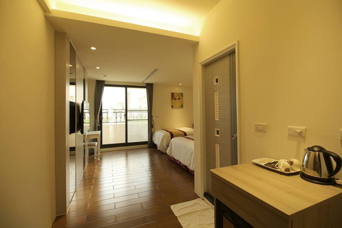 Habitación de hotel con piso de madera, una cama, un escritorio con hervidor y una ventana con cortinas.