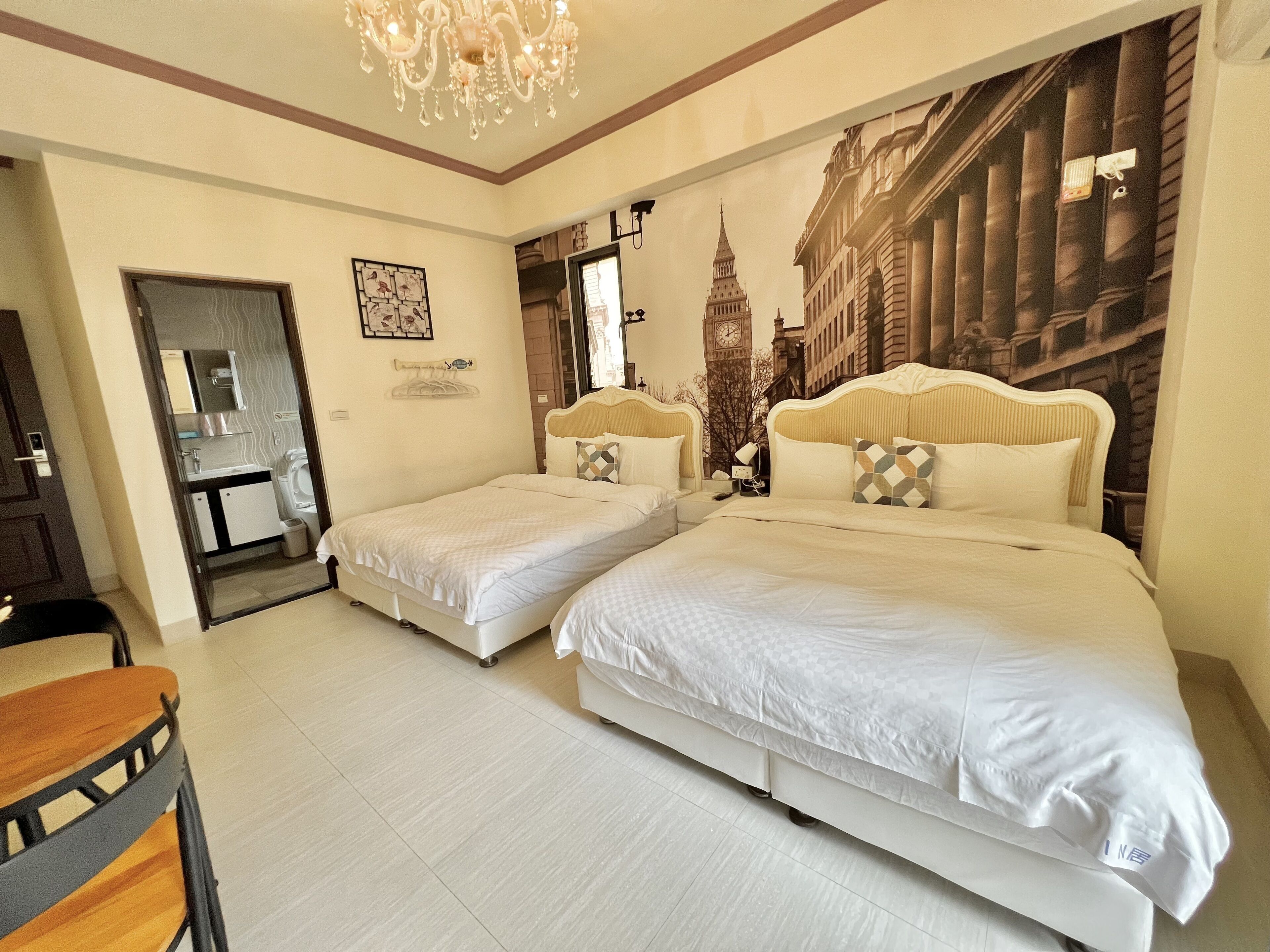 A hotel room with two beds, a chandelier, a bathroom with a mirror, and a wall mural of a cityscape.