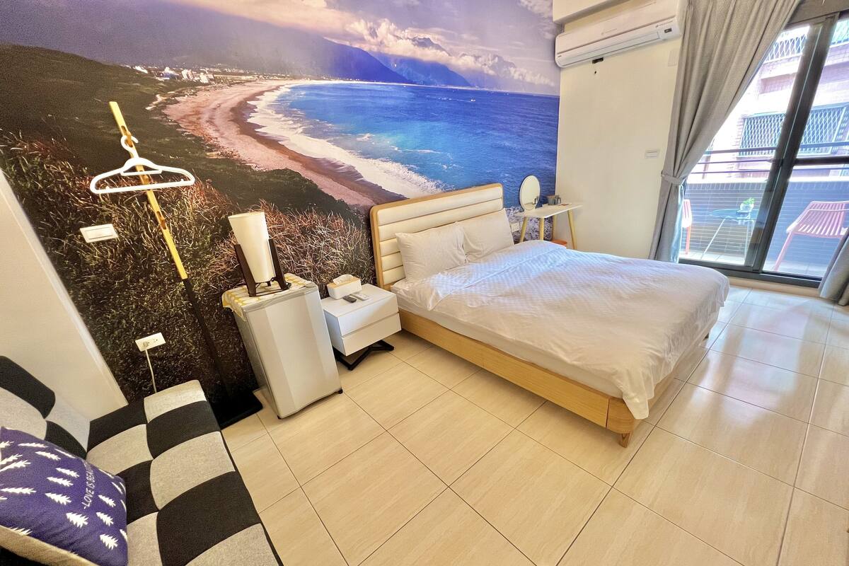 A hotel room with a bed, nightstand, and a wall mural of a beach scene.