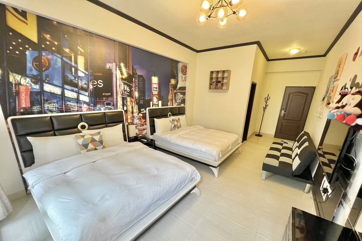 A hotel room with two beds, a large window with cityscape view, a chandelier, and a small shelf with items.