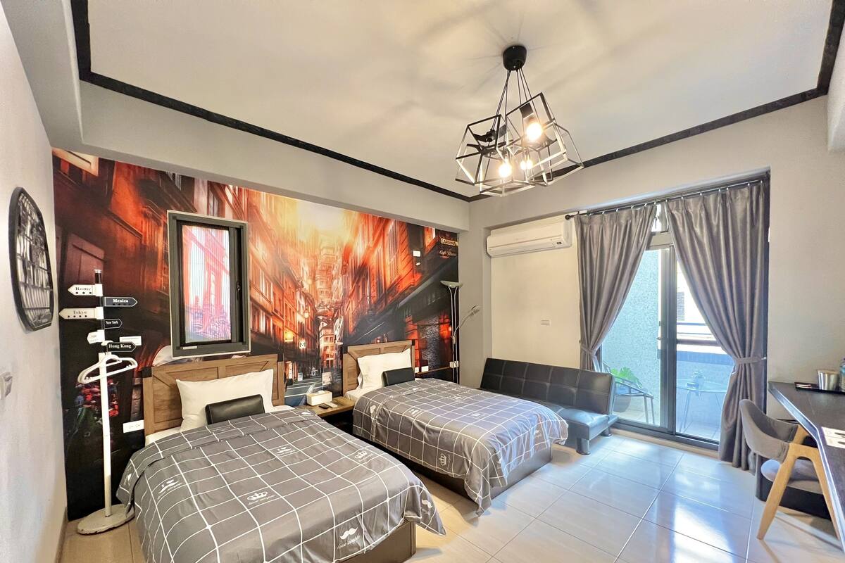 A hotel room with two beds, a desk, a chair, a ceiling light, and a large window with curtains.