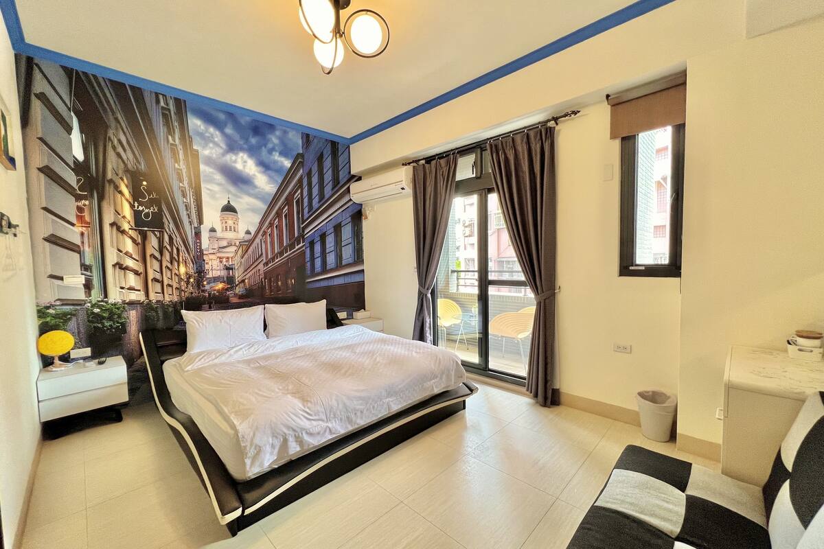 A hotel room with a bed, a desk, a chair, a window with curtains, and a view of buildings outside.