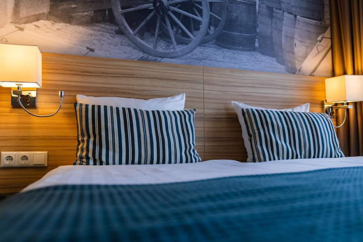 Une chambre d’hôtel avec une tête de lit en bois, deux coussins à rayures et des lampes fixées au mur.