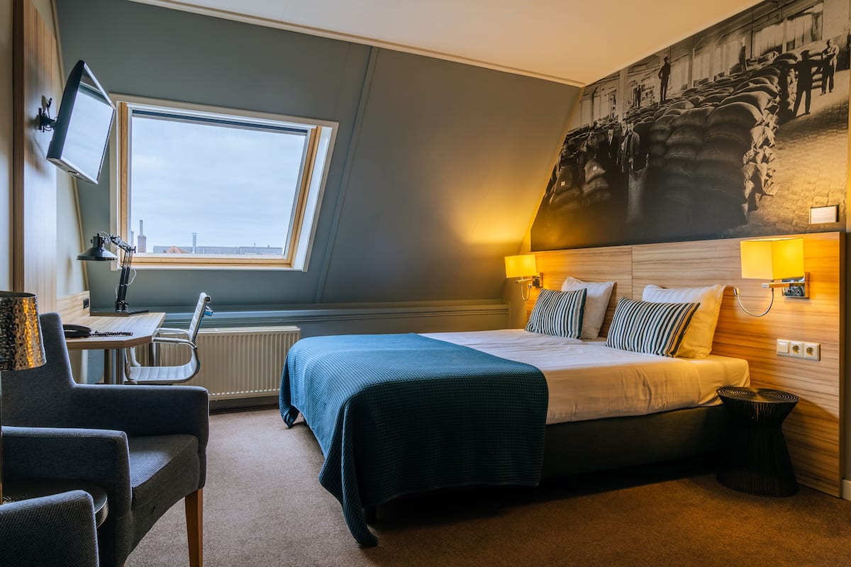 Une chambre d’hôtel comprenant un lit, des lampes de chevet, un bureau avec une chaise, une télévision et une grande fresque murale.