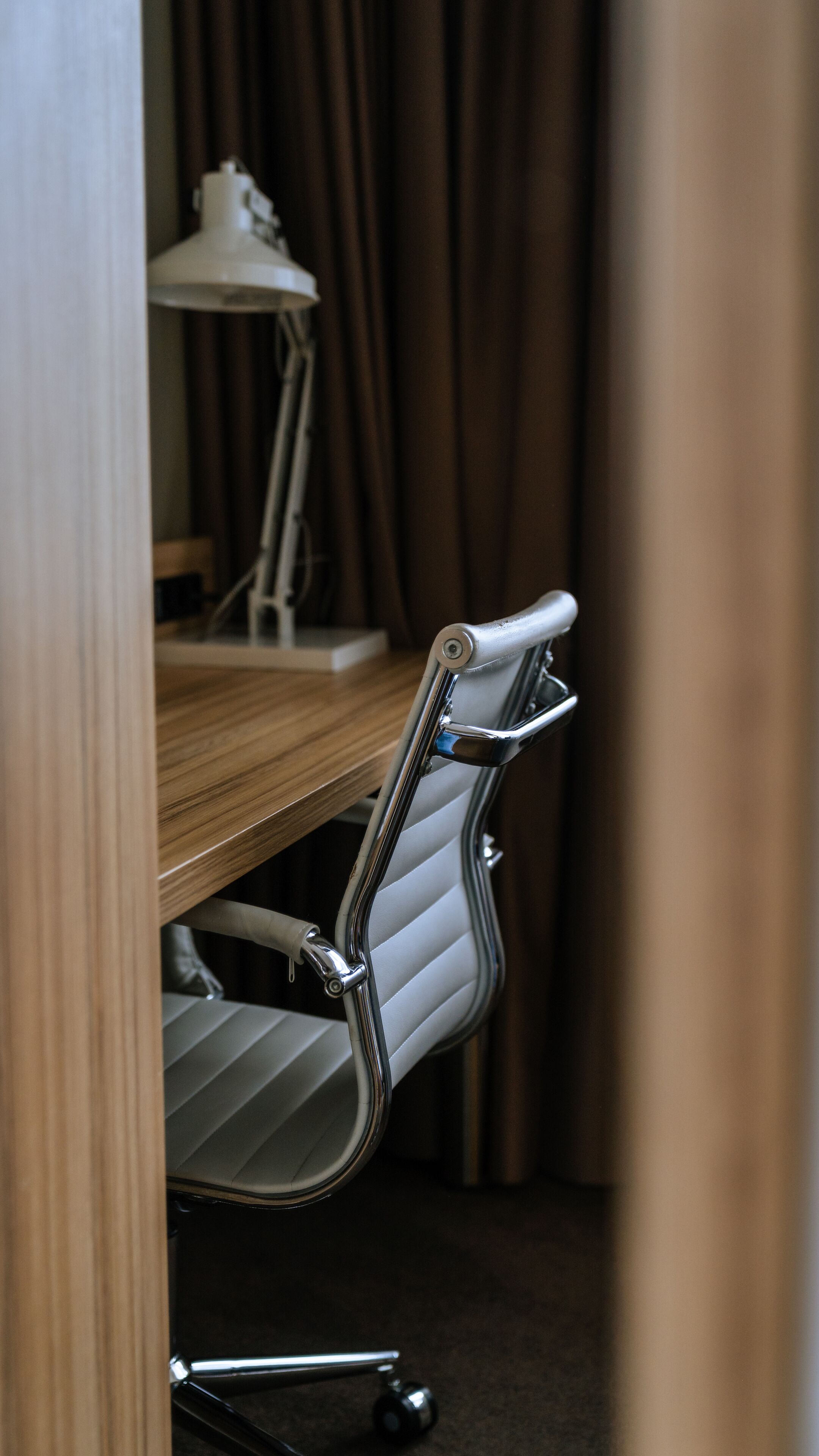 A modern office chair with a metallic frame and white upholstery, positioned at a wooden desk with a white lamp in the background.