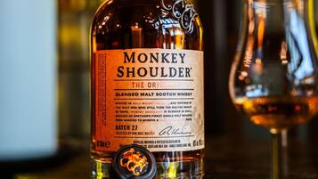 Una botella de whisky Monkey Shoulder sobre una barra con un vaso y una vela al fondo.