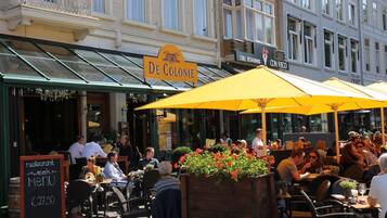 Un café al aire libre con sombrillas amarillas, un letrero que dice 'De Colonie' y un cartel de menú para la 'Semana del Restaurante'.