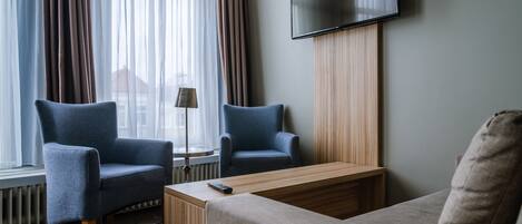 A hotel room with two blue armchairs, a wooden coffee table, a grey sofa, and a flat-screen TV mounted on the wall.