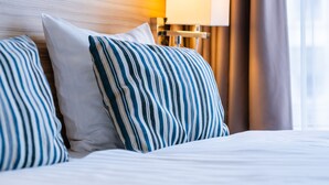 Une chambre d’hôtel avec un lit, des coussins à rayures, une lampe et une grande fenêtre avec des rideaux.