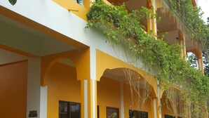 A two-story building with yellow and white walls, a balcony with blue balustrades, and a well-maintained garden with a hedge and potted plants. - Mood Mansion (Kamala)