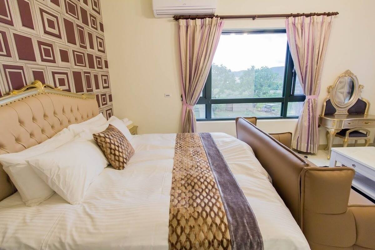 Habitación de hotel con una cama grande, cabecera, mesita de noche, espejo y ventana con cortinas.