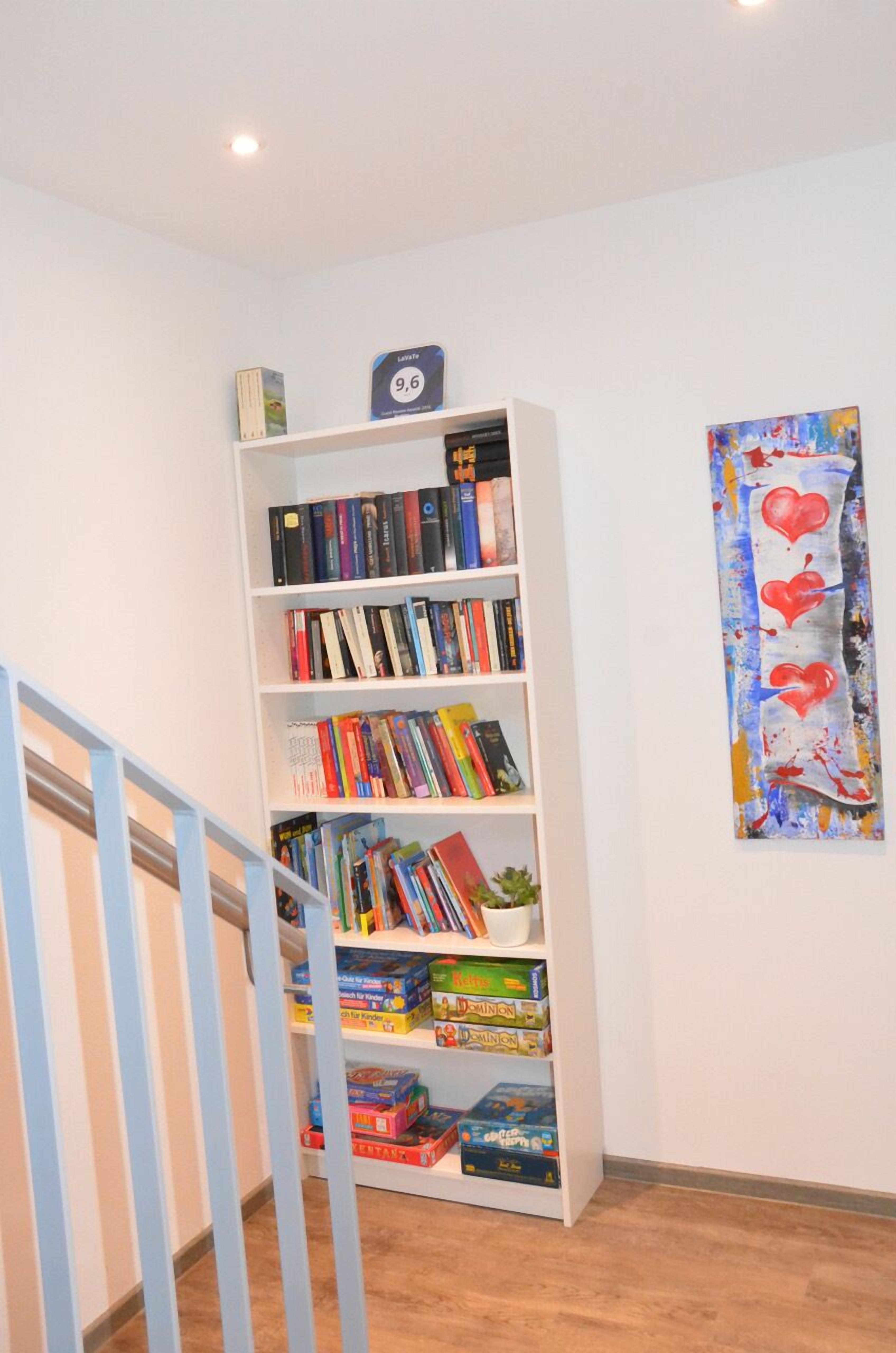 A bookshelf with books and board games, a painting on the wall, and a staircase.