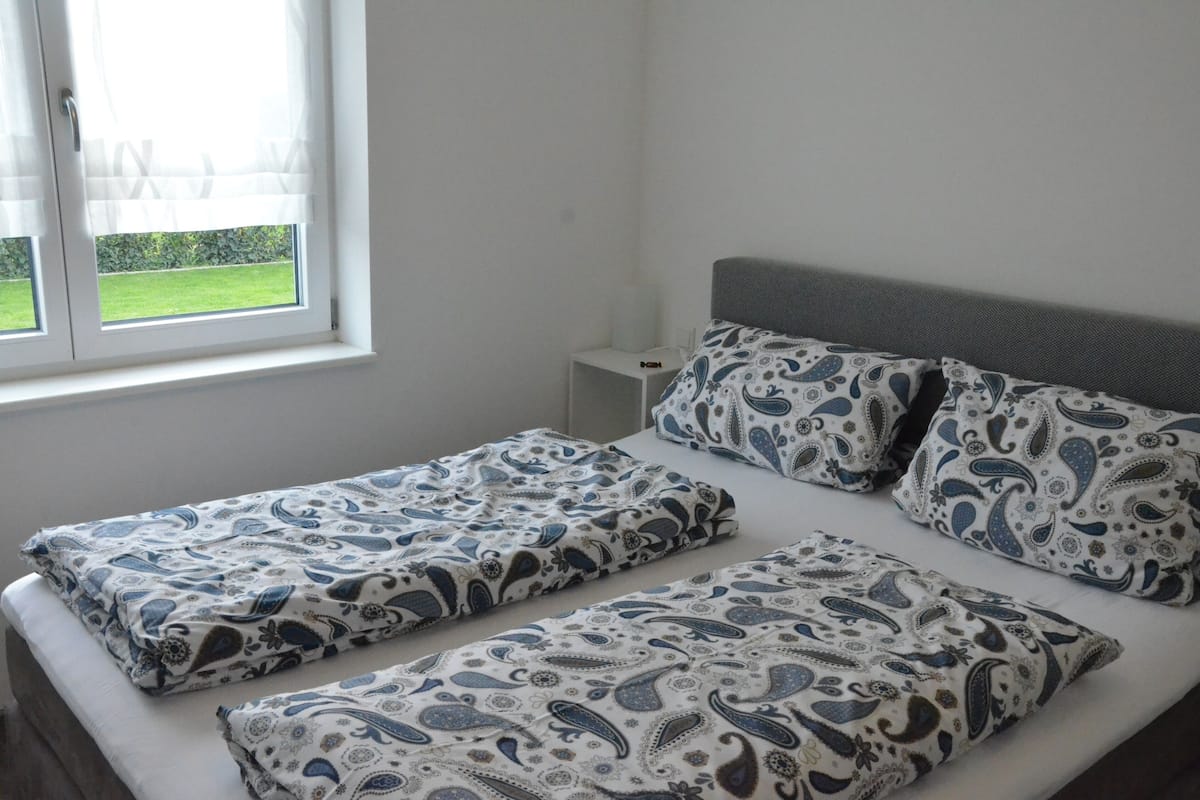 A double bed with matching patterned bedding, a bedside table, and a window with a view of greenery.