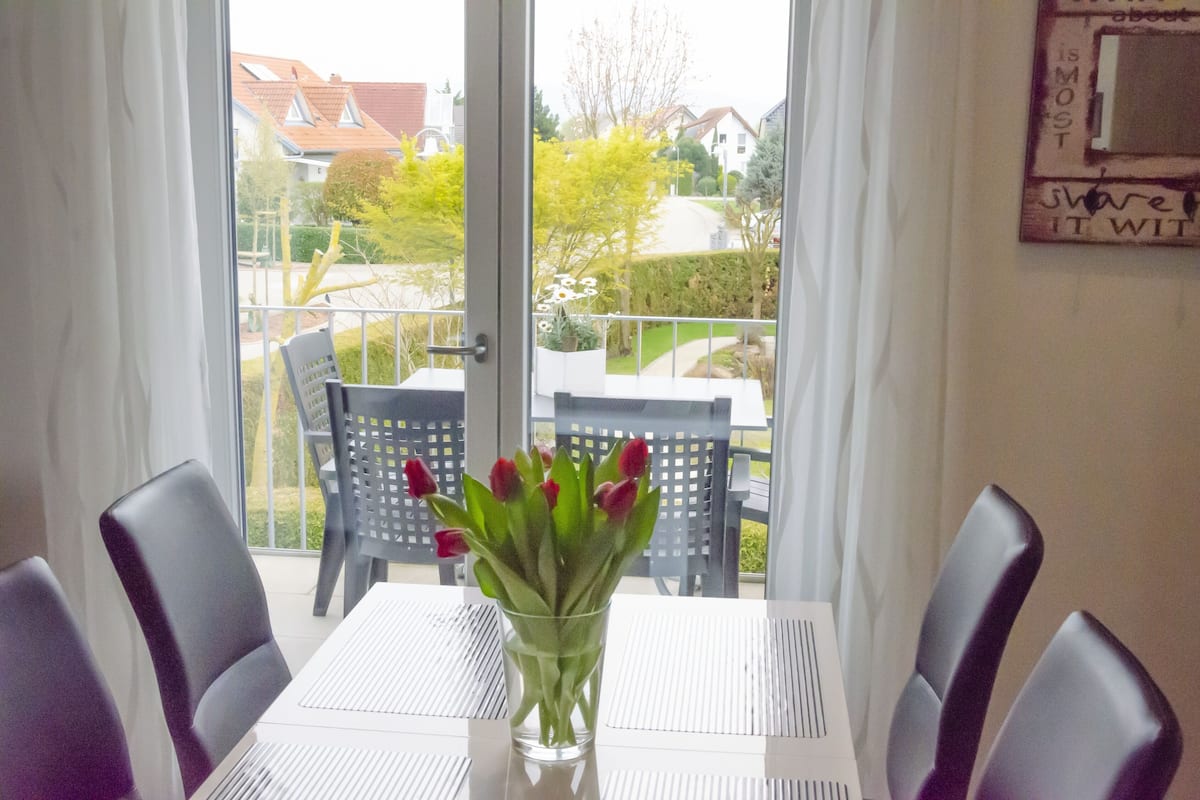 A dining area with a table and chairs, a vase with red flowers, and a view of the outside through a glass door.