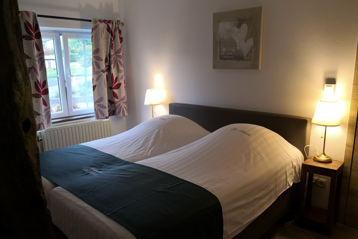 A neatly made bed with white linens and a green blanket, two bedside lamps, a window with floral curtains, and a wall-mounted artwork.