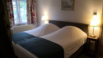 En pænt redt seng med hvide sengetøj og et grønt tæppe, to sengeborde med lamper, et vindue med blomstrede gardiner og et billede hængt på væggen.