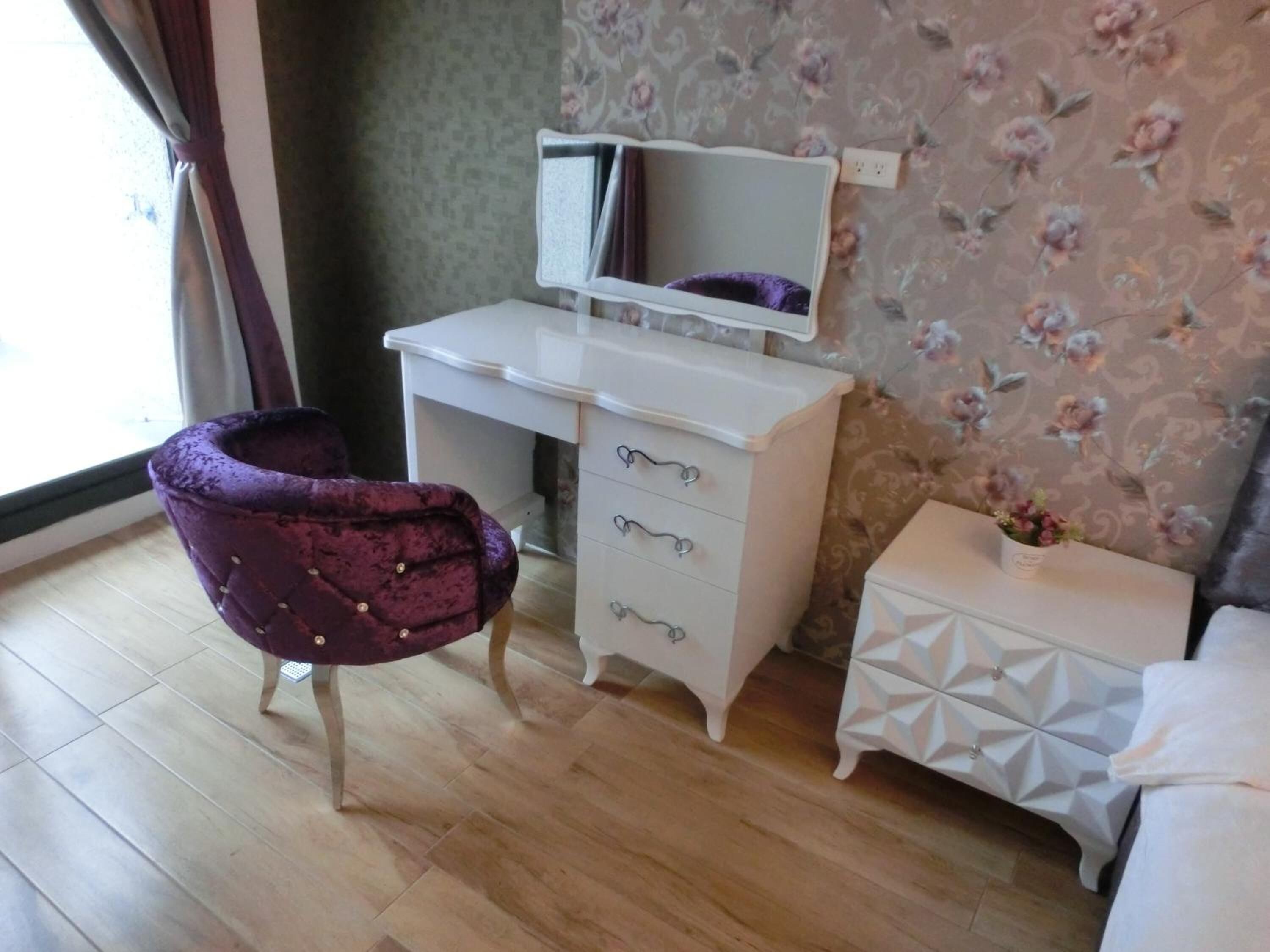 Une chambre avec un plancher en bois, un bureau blanc avec des tiroirs, une chaise violette, une table de chevet blanche ornée d’un vase de fleurs et un lit.