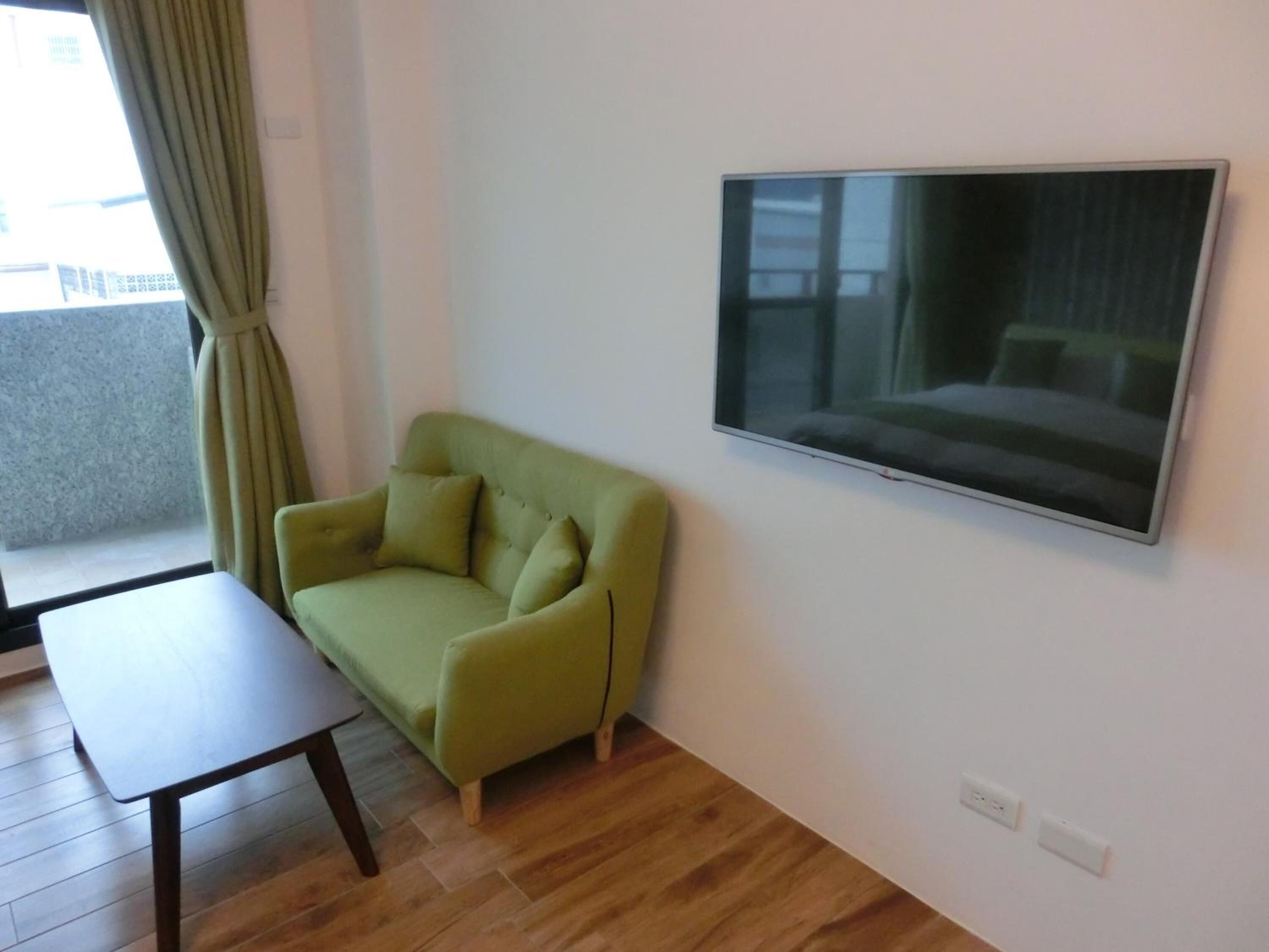 Una habitación con un sofá verde, una mesita pequeña y un televisor instalado en la pared.