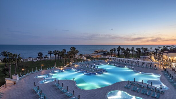 A resort pool area with multiple swimming pools, lounge chairs, and a view of the ocean at dusk. - Arcanus Hotels Sorgun - All Inclusive (Manavgat)