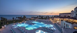 A resort pool area with multiple swimming pools, lounge chairs, and a view of the ocean at dusk.