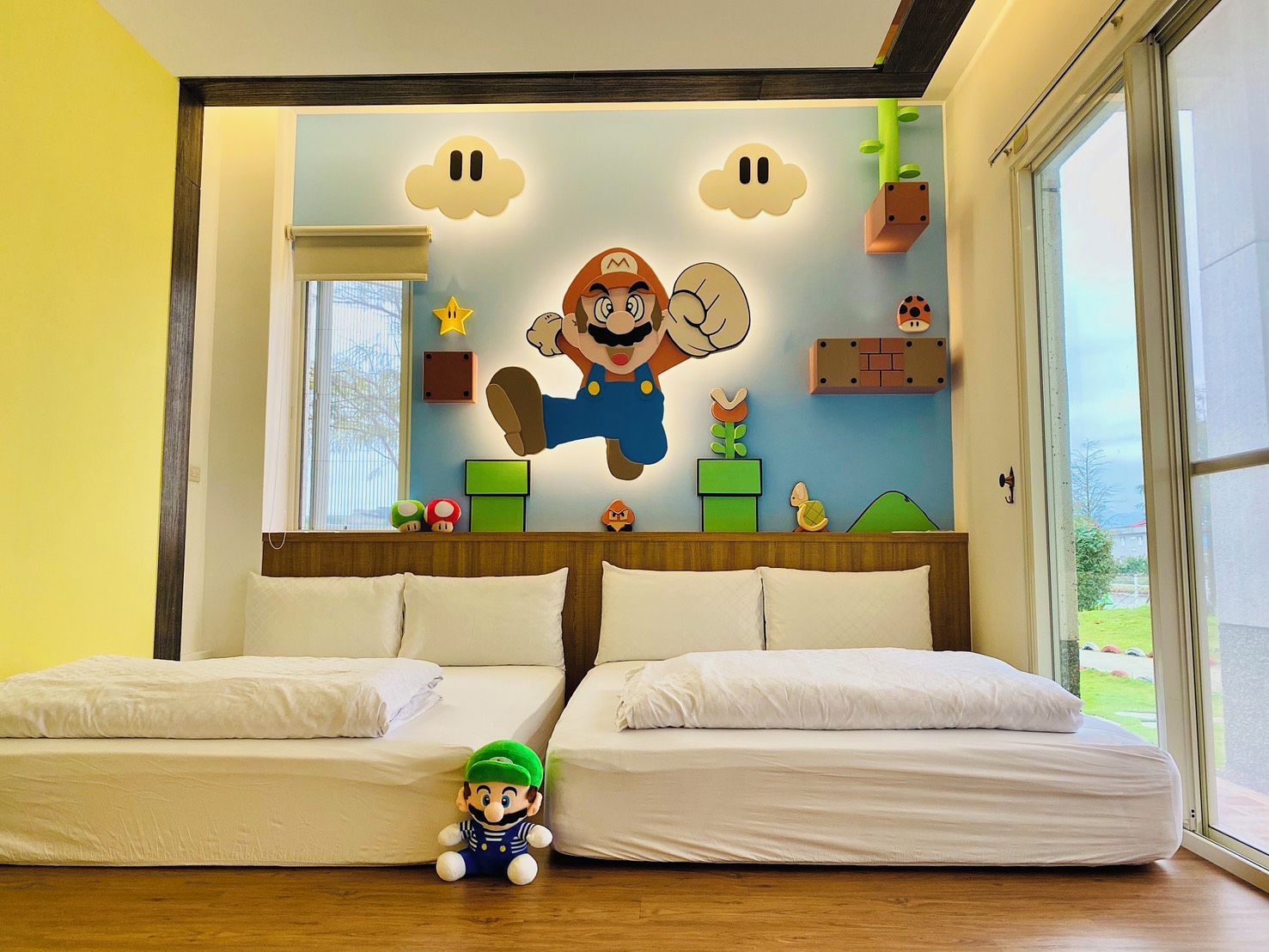 A room with two beds, a large window, and a wall decorated with Mario and Luigi characters.