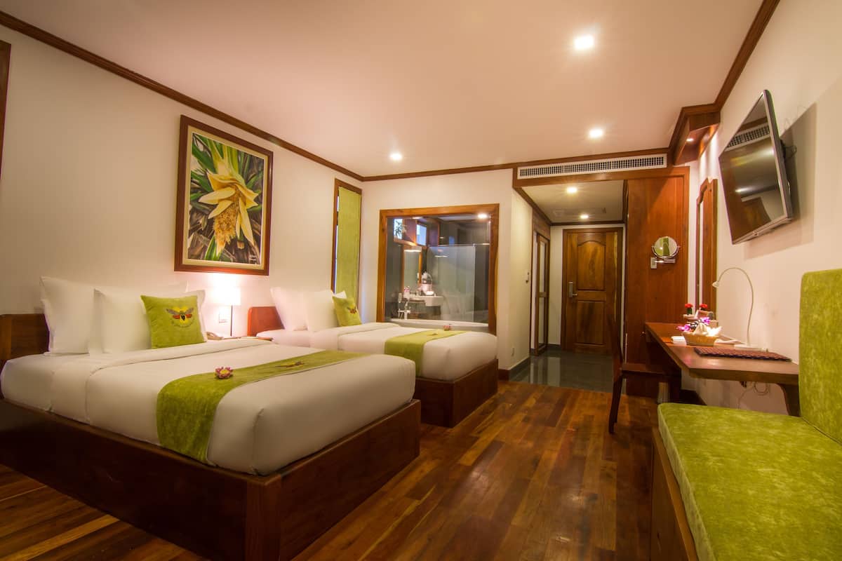 Habitación de hotel con dos camas, un escritorio, una silla, un televisor y un baño visible a través de una puerta de cristal.