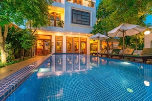 A swimming pool with a tiled edge, surrounded by lounge chairs and umbrellas, and a building with multiple windows and balconies.