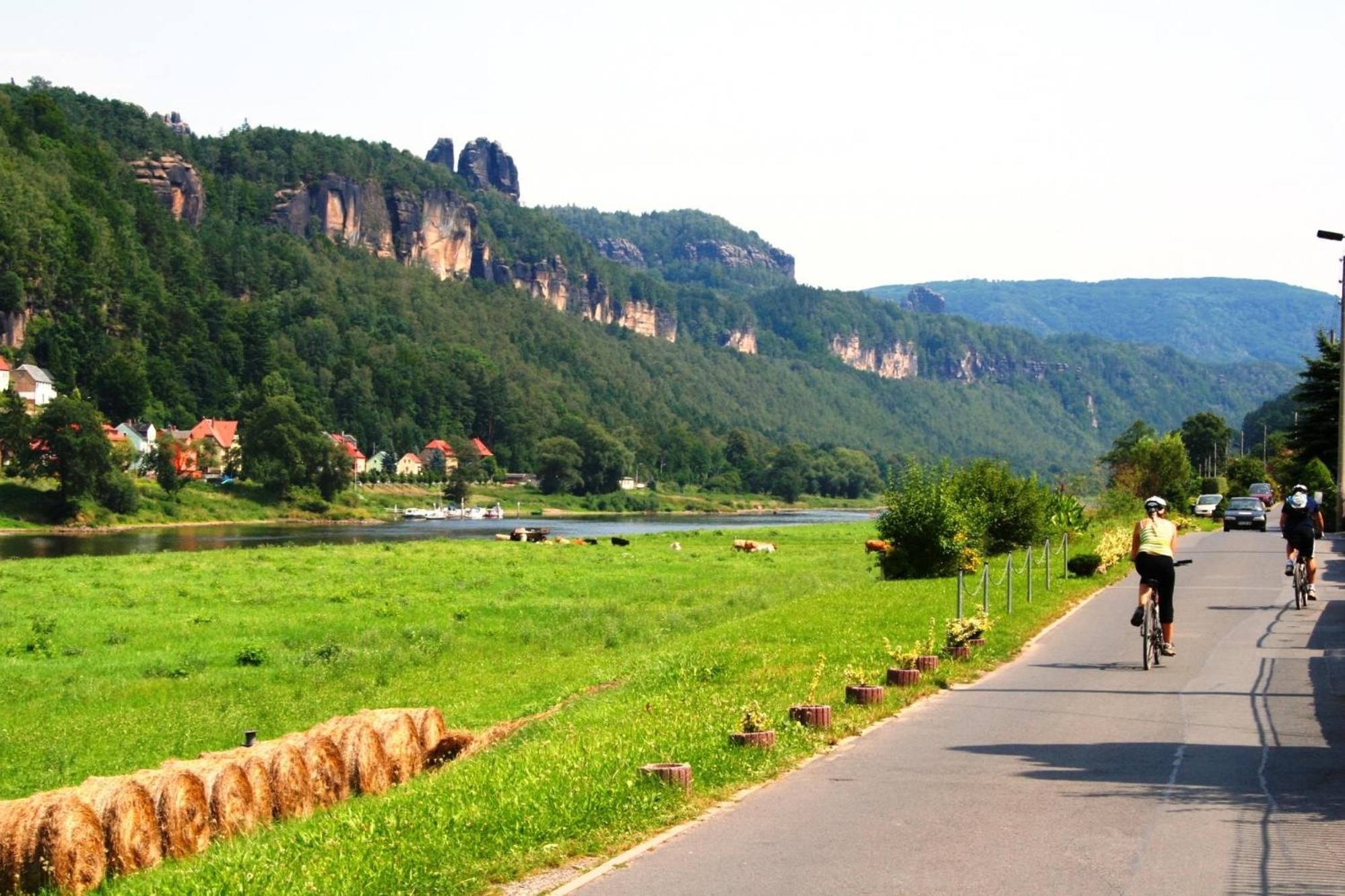 A paved road with cyclists, a river, and a forested hillside with houses.