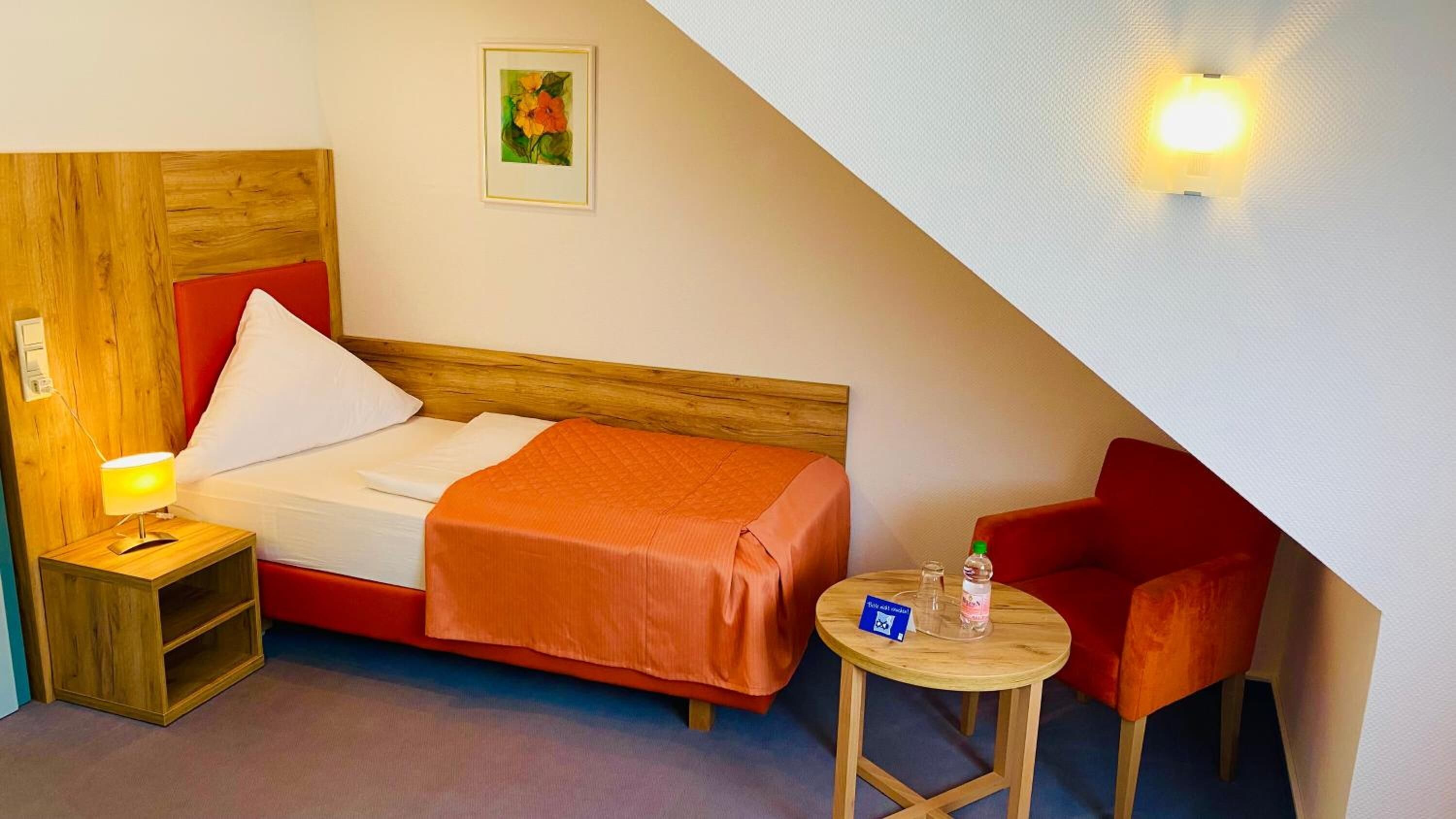 A hotel room with a bed, a nightstand, a red chair, a small table, and a wall-mounted light.