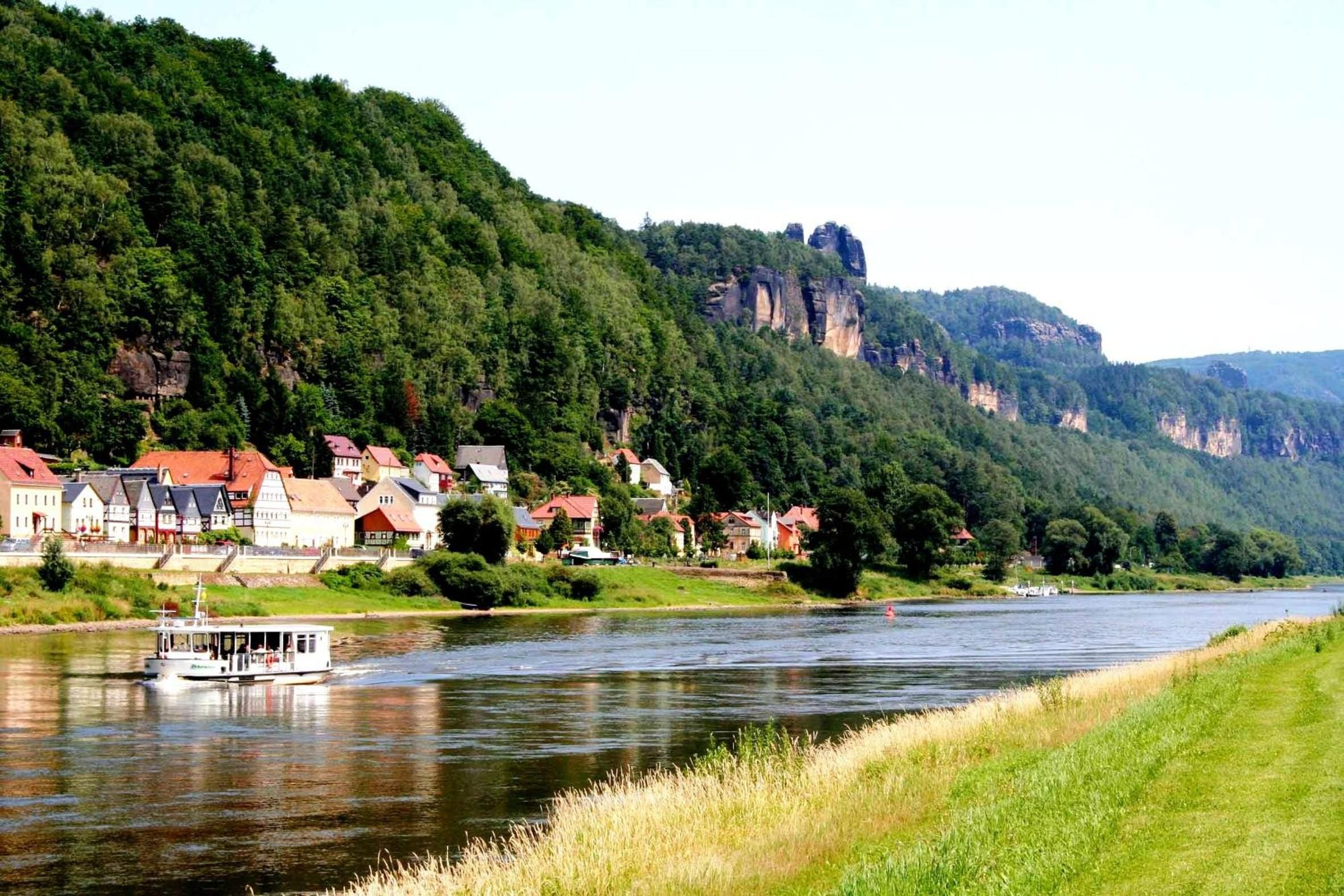A river with a boat, houses along the riverbank, and a forested hill with a prominent rock formation.