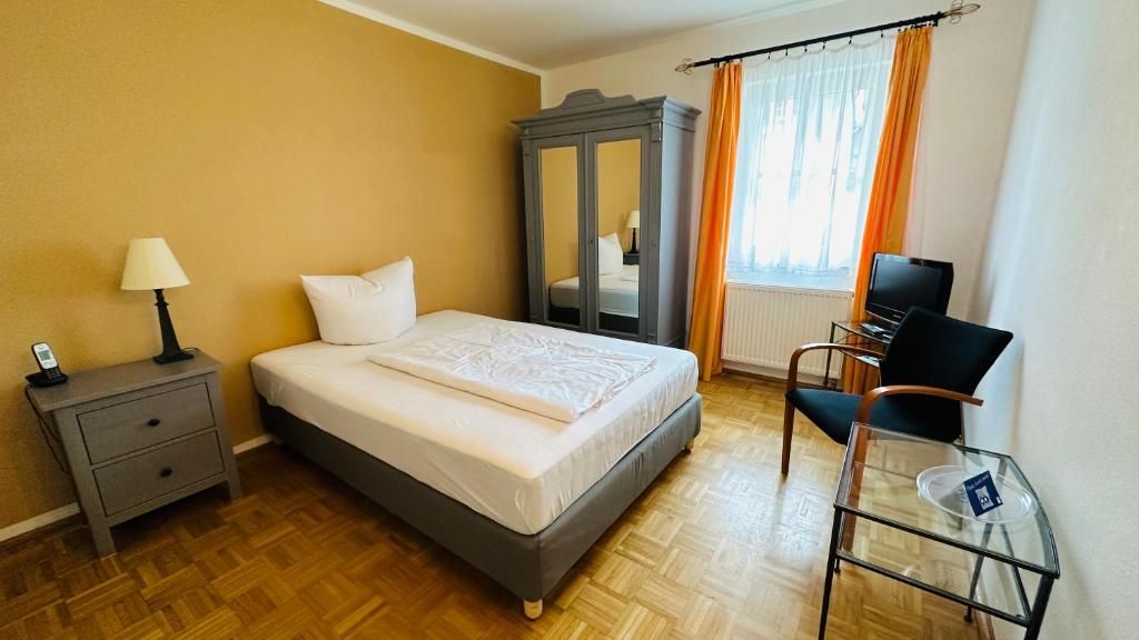 A hotel room with a bed, a nightstand, a lamp, a wardrobe, a chair, and a glass table.