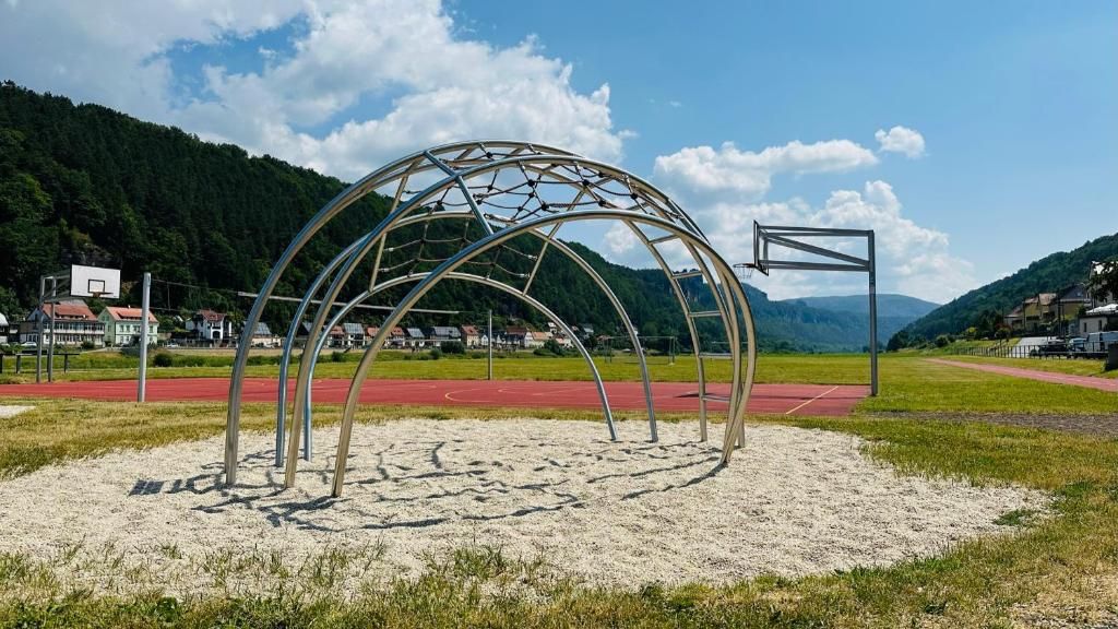 A metal structure resembling a tunnel or arch in a park setting with a red track and houses in the background.