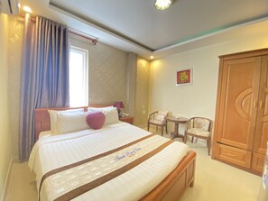 Una habitación de hotel con una cama grande, un armario de madera, dos sillas, una mesita y una ventana con cortinas.