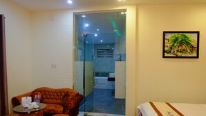 A hotel room with a bed, a sofa, a small table with items on it, and a glass door leading to a bathroom.