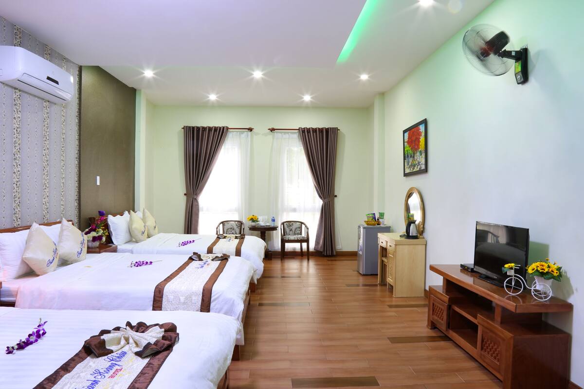 A hotel room with four beds, a TV, a fan, a small table with a vase of flowers, and a bicycle on a wooden stand.