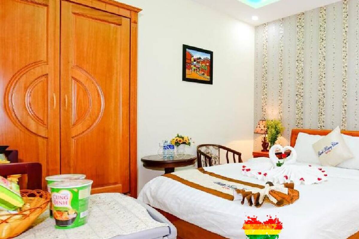 A hotel room with a bed, a wooden wardrobe, a small table with a lamp, and a chair.