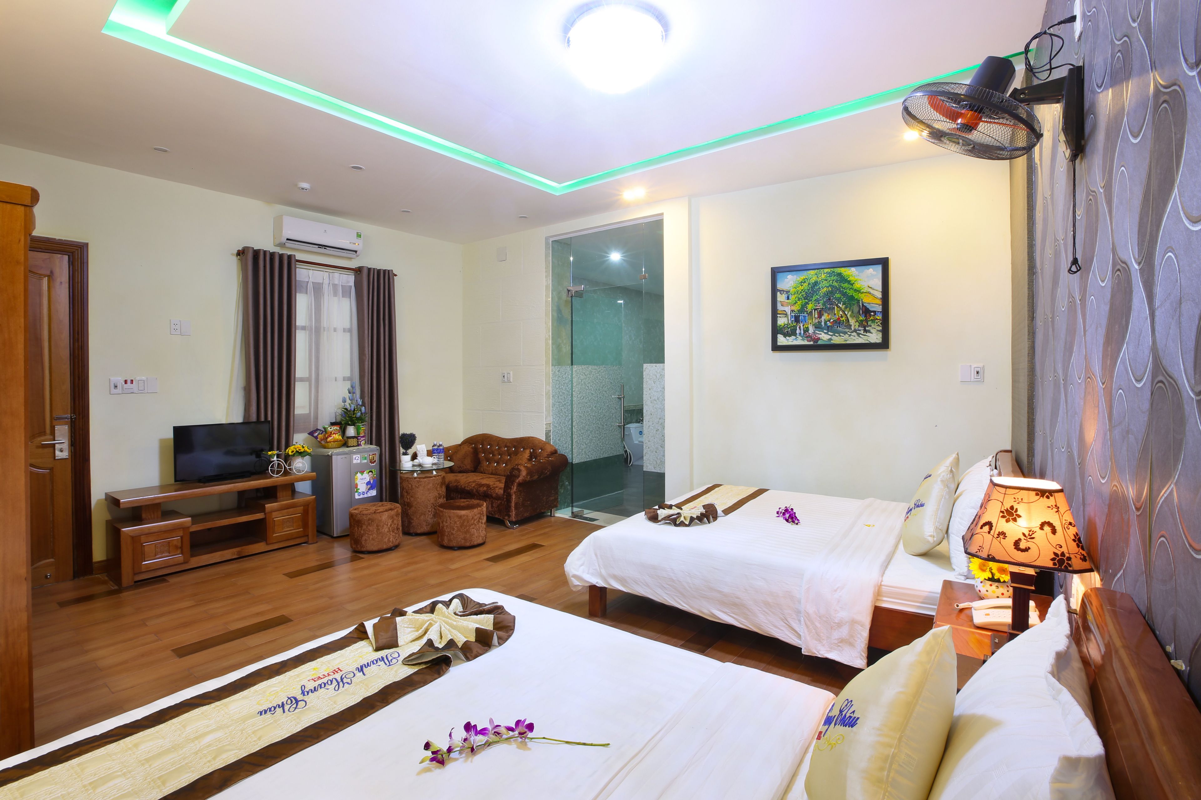 A hotel room with two beds, a TV, a sofa, a small table, and a wall-mounted fan.