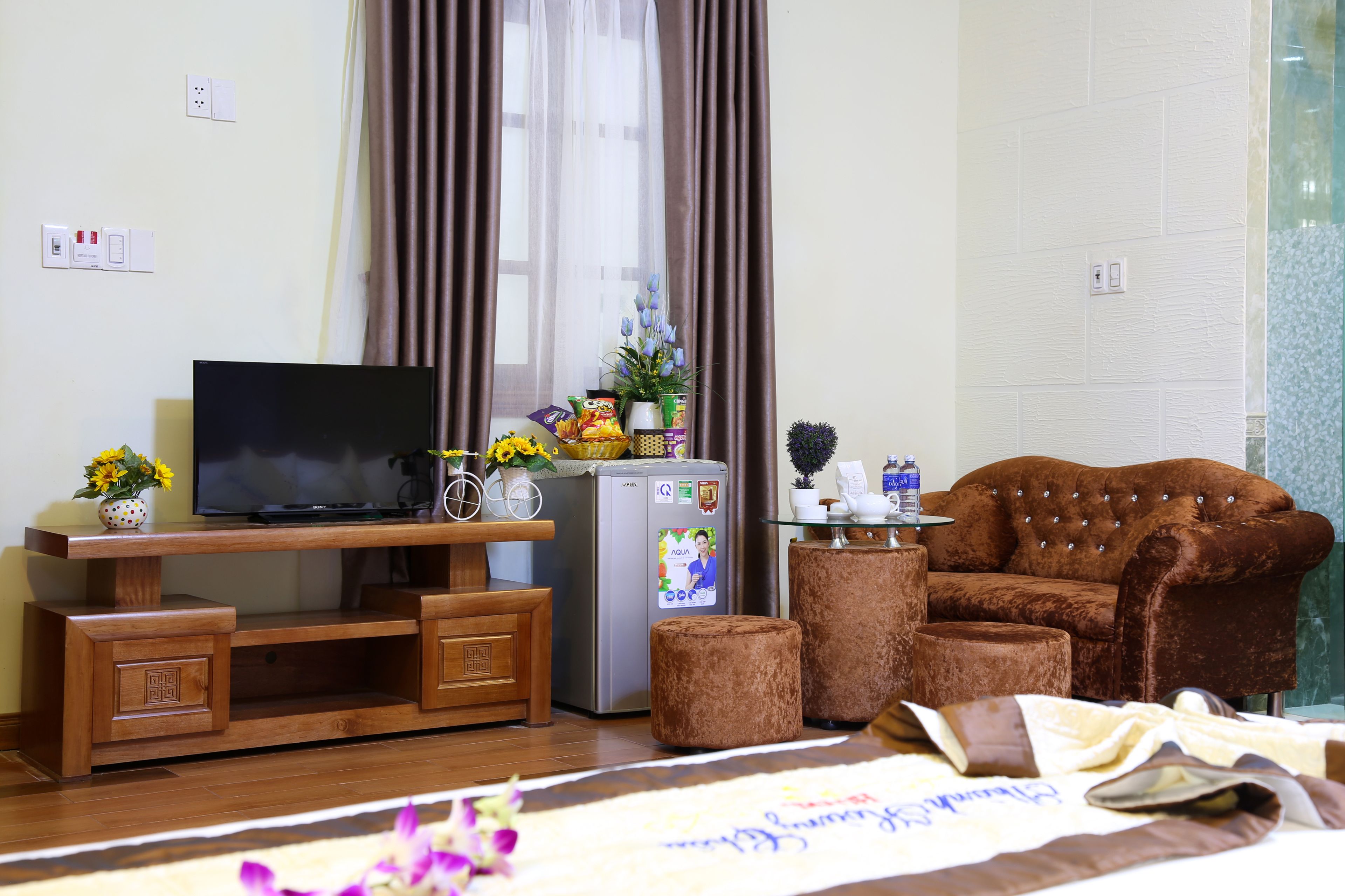 A living room with a TV, wooden entertainment unit, refrigerator, brown sofa, and ottoman.