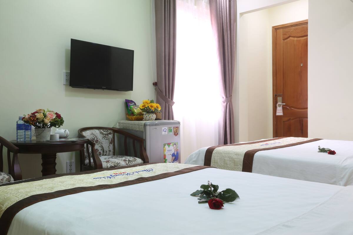 A hotel room with two beds, a TV, a refrigerator, a table with flowers, and a door.
