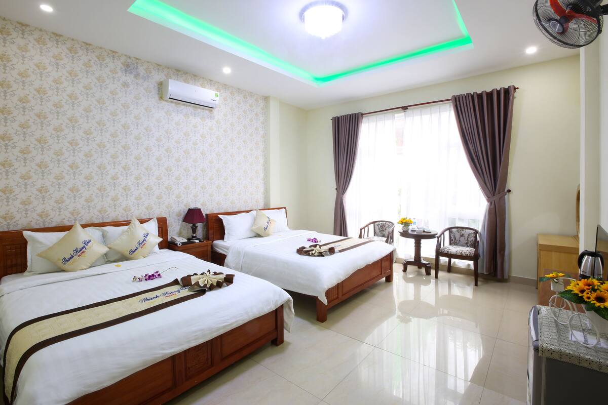 A hotel room with two beds, a small table, a chair, a fan, and a window with curtains.