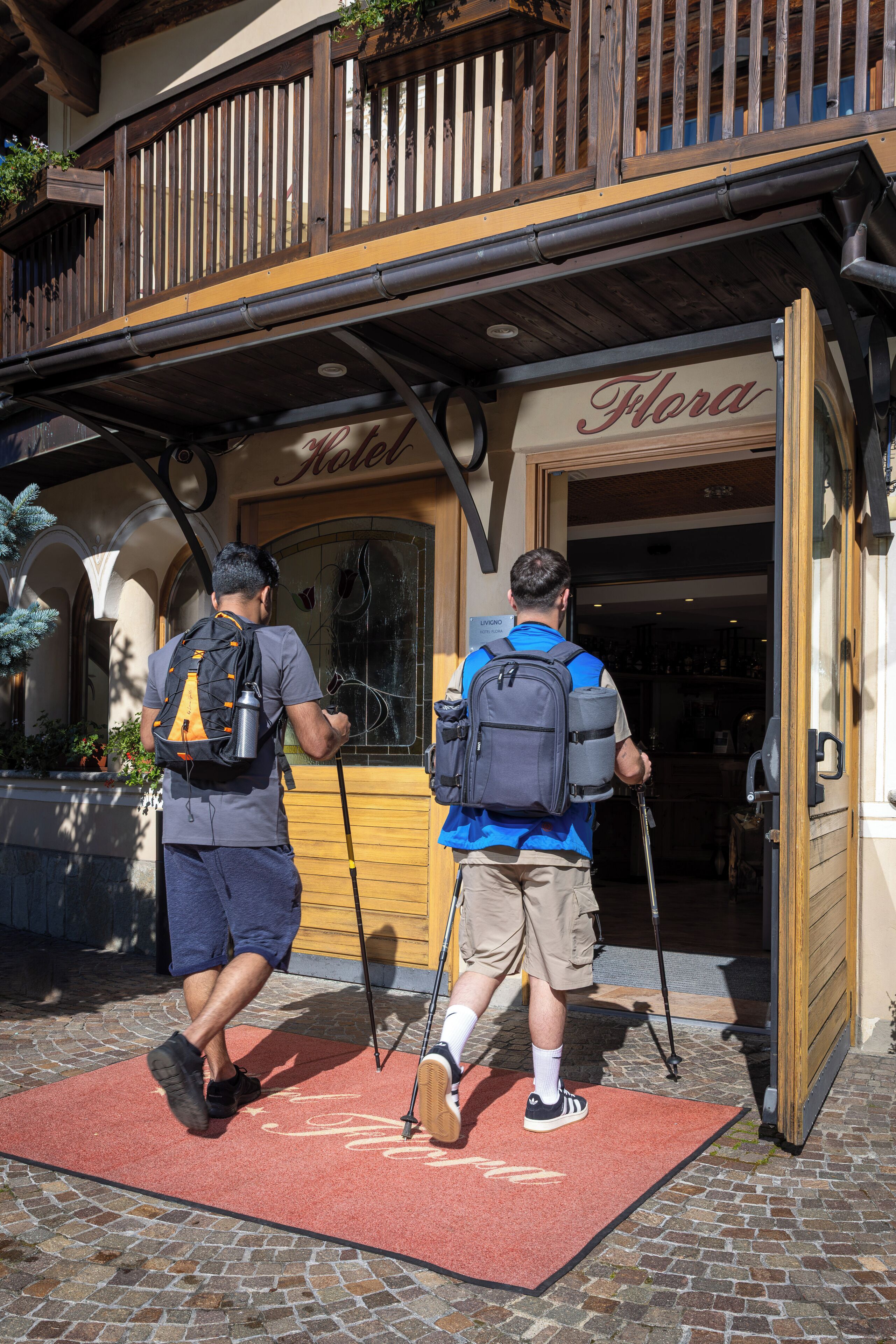 Two people entering a hotel with a wooden balcony and a red doormat.