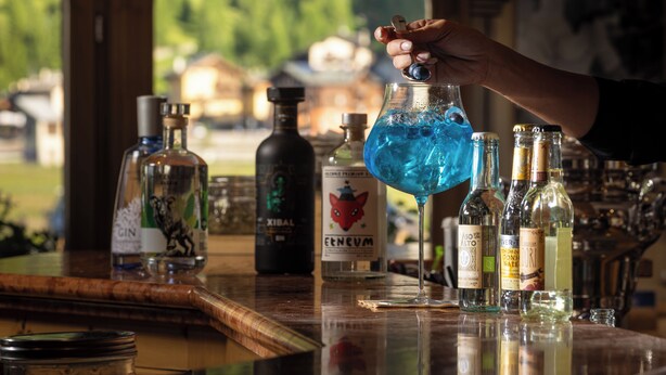 A bar with various bottles and a hand pouring a blue liquid into a glass. - Hotel Flora (Livigno)