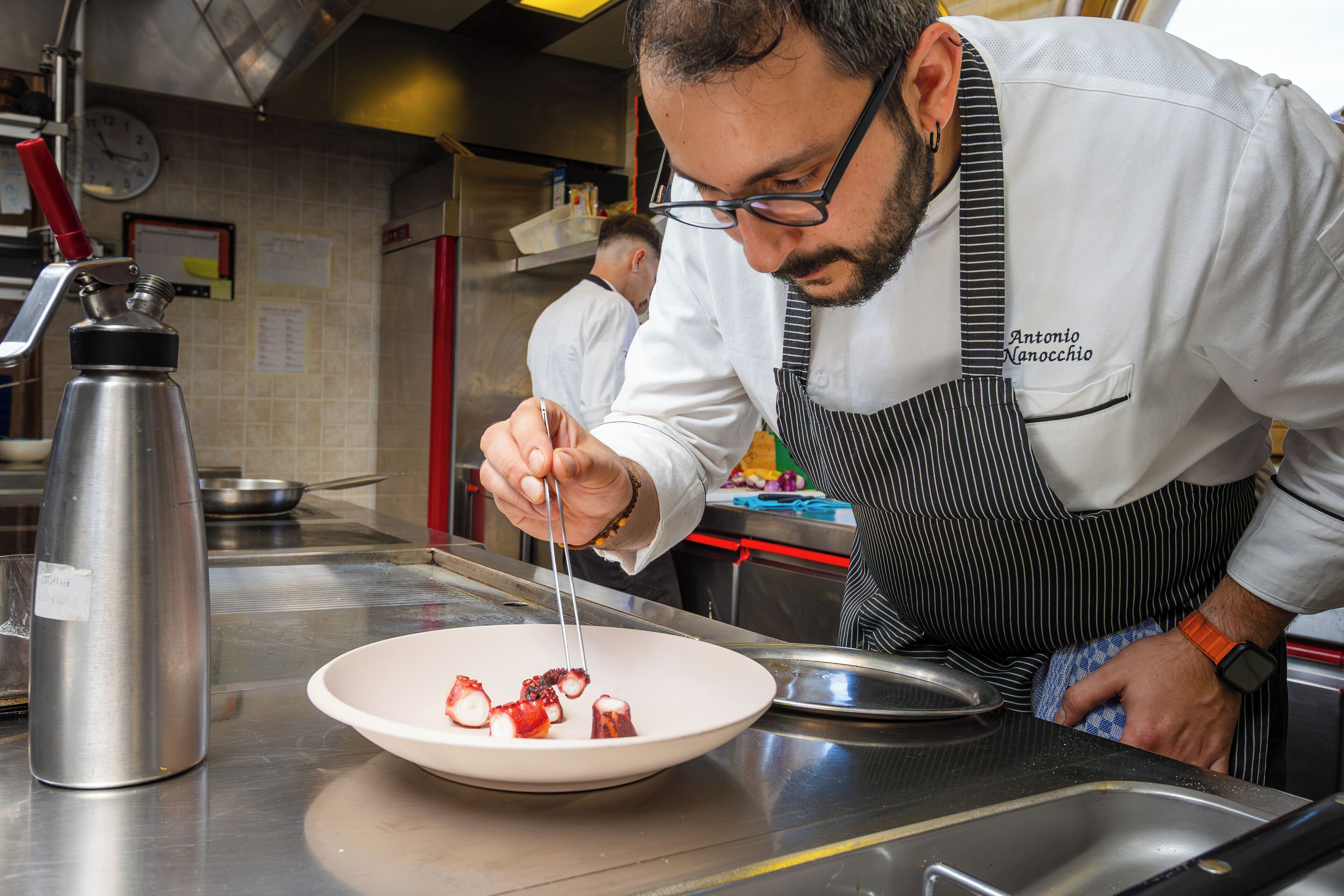 A chef preparing food in a kitchen with stainless steel appliances and a clock on the wall.