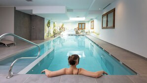 A woman relaxing at the edge of an indoor swimming pool with a view of a mural and a shower area. - Hotel Flora (Livigno)
